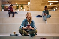 Das Bild zeigt eine Studentin, die an der Uni sitzt und in ihr Handy schaut. 
