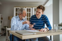 Das Bild zeigt einen älteren Mann, der mit einem jungen Mann an einem Tisch sitzt. 