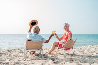 Das Bild zeigt einen älterer Mann und eine ältere Frau, die in Liegestühlen am Strand sitzen und anstoßen.